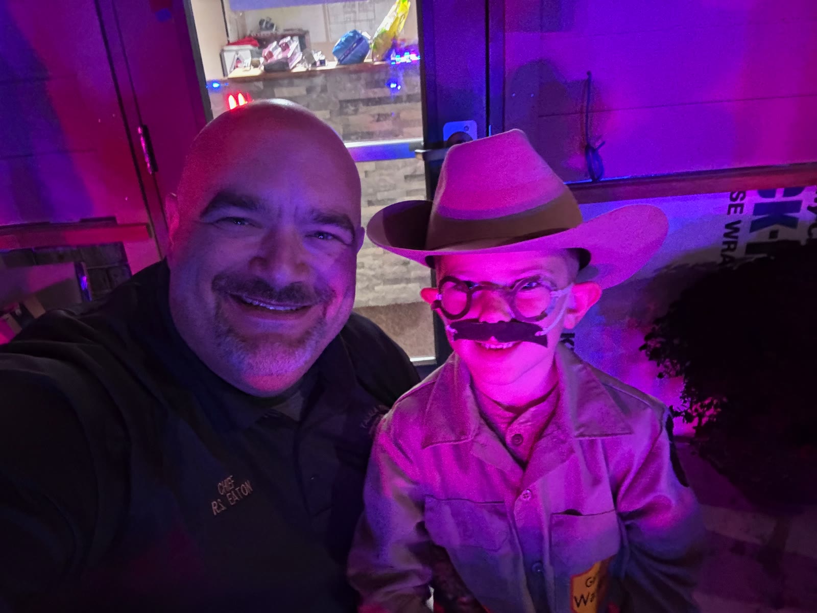 Chief R. Eaton selfie with a smiling young boy in a cowboy hat and costume mustache under purple event lighting