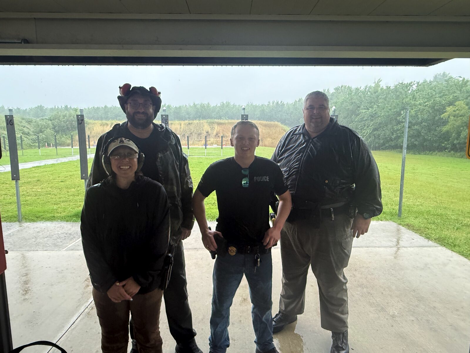 Talala PD officers and partners under a covered firing-range bay on a rainy day, with wet range lanes and markers in the background
